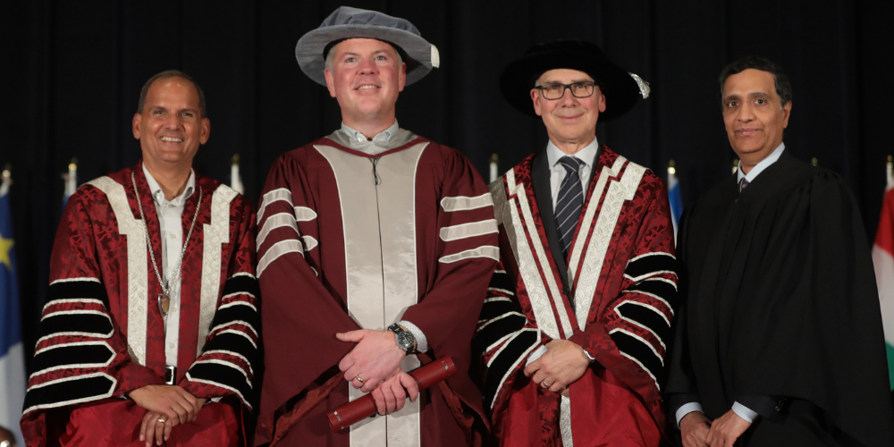 Jonny stands on stage at convocation with the President, Chancellor and a professor