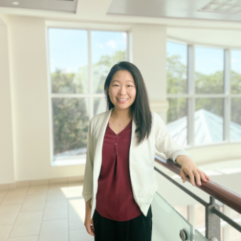 Wendy stands with her hand resting on a railing inside a sunny room. She has olive skin,  shoulder-length dark hair and wears a red blouse, white blazer and black pants.